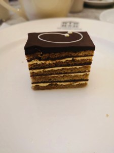 A slice of signature gateau on a white plate. The gateau is made from many thin layers of sponge, cream and chocolate 