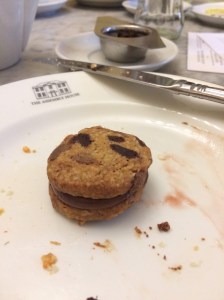 Chocolate chip cookie sandwich with chocolate cream in the centre. On a white plate with crumbs and food stains. You can see a Tea strainer in the distance
