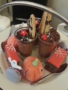 Looking down at the top tier of a cake stand onto sweet treats. There is a pumpkin profiterole, a small glass with a chocolate mousse and RIP headstone, a macaron with a ghost made of icing. 