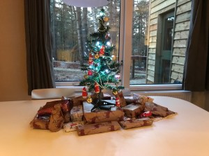 A mini Christmas tree on the white dining room table. There are small gifts wrapped up under the tree. The window behind looks out onto trees and a pathway. 