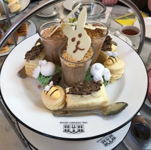 Looking down onto the top tier of the afternoon tea. There is a yellow layered gateau style cake, a yellow macaron with a bumble bee made of icing on top, a mini cupcake that has a white rabbits bum and legs sticking out made of icing, and a small glass filled with mousse topped with a which chocolate bunny face. 