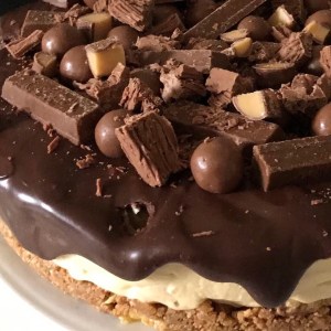 A large cheesecake covered in chocolate ganache and broken up chocolate including kit kats, malteasers, and fudge.