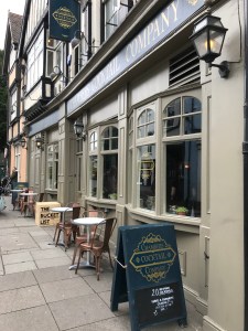 Looking down the street there is Chambers Cocktail Company on the right. Tables are outside and a wooden cube has the words The Bucket List on outside to doorway