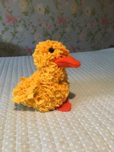 A bright yellow fluffy duck made of felt, it appears to have tufts. It has an Orange felt beak. It is sitting on my light blue with white spots bed cover. 