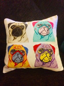Four cross stitch pugs in pop art style in various colours. One pug is natural colours, one is bright yellow and red, one is a turquoise/mint colour with pink, and one is purple and yellow. 