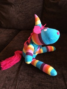 Photo of the sock unicorn taken from the side. He is stuffed and sitting up. He has a pink wool tail and pink ears. 