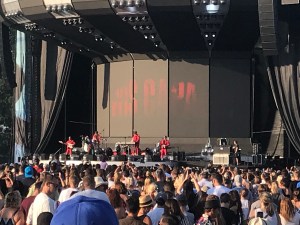 Lewis Capaldi is on stage with his band all wearing red. Lewis is in a bright multicoloured tracksuit