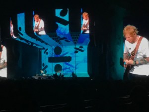 Ed Sheeran is on stage. It’s blue, with the Divide logo as the backdrop, and he can be seen on the large screens. 