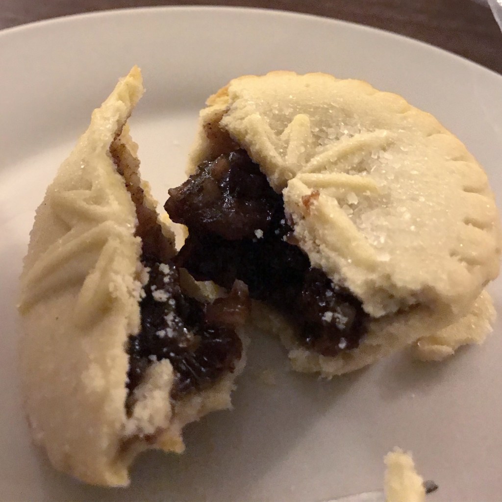 A crisp mince pie with a pastry star on top. The mine pie is broken in half so that you can see the filling coming out. It is on a while plate