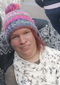 A headshot of myself, selfie style. I am wearing a stripy rainbow multicoloured knitted hat, and a cream coloured scarf with small birds printed on.