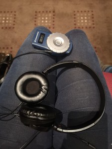 A photo taken looking on to Lucy’s lap. She is wearing blue jeans. The headphones and audio description box are laying on her lap. 