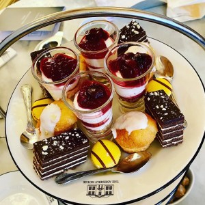 A photo taken looking down onto the top tier of a cake stand. There is a shot glass filled with cherry cheesecake, a chocolate layered sponge cake, lemon drizzle cupcake, and yellow banana flavour macaron. 
