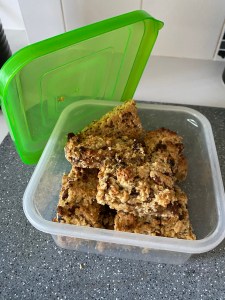 A plastic container with homemade flapjacks in. There are sultanas in the bakes. The container has a green lid which is open and propped to the side. 