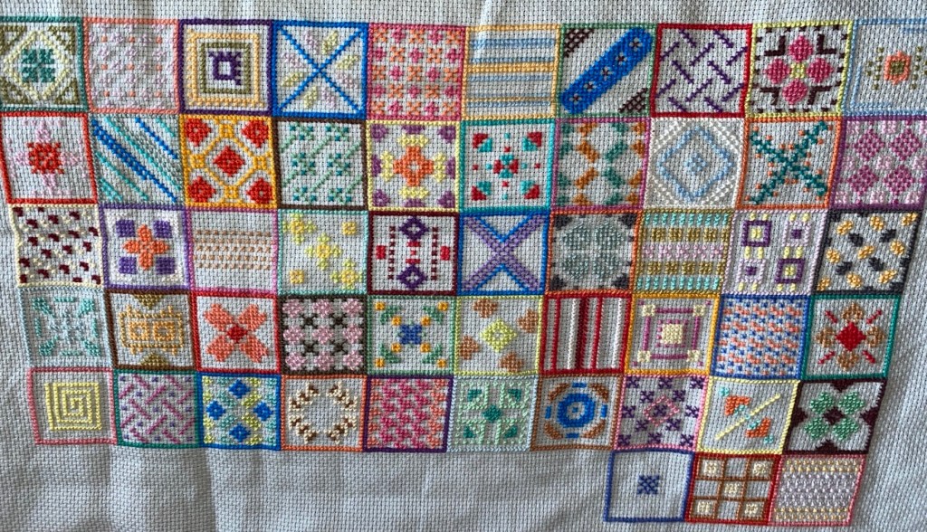 This photo is taken at a distance from the cross stitch fabric. All the squares are in rows of 10 each one touching the next. Like a grid filled with patterns. A variety of every colour is used and each design is different. 
