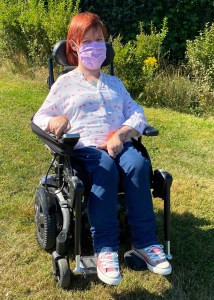 A photo of me in my power wheelchair. I have blue jeans, a white shirt with pink flamingos on, I have red hair and am wearing a light purple homemade fabric face mask. I am on grass and there are plants in the background. 