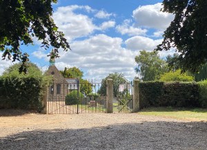 Iron gates lead into the graveyard with trees either side. The chapel is the the left and the sky is blue and sunny with white clouds. 