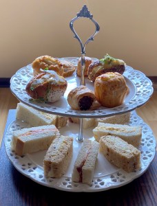 A two tiered cake stand with finger sandwiches on the bottom, and savoury bites on the top. Including a roll, a biscuit, a sausage roll and a muffin. 