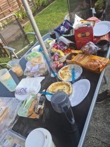 A black glass garden table with parasol pole is scattered with paper plates and packets of food including sausage rolls, quiche and cups of drink. 
