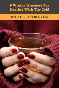 An image to pin. Title text 9 Winter Warmers For Dealing With The Cold, is at the top in orange text on dark grey background. The below image is a close up of hands holding a mug, they have red painted nails and red knitted sleeves showing.