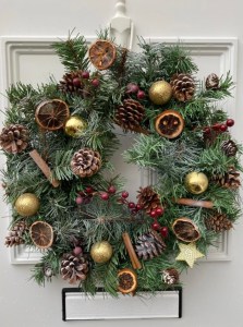 The second wreath is hanging on a white door. There is a letter box at the bottom. The wreath is green with gold baubles, cinnamon sticks, orange slices, pine cone and red berries. 