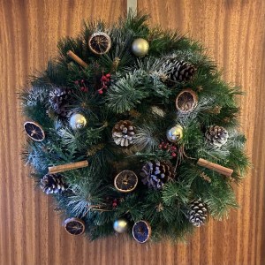 The wreath I constructed is hanging on a wooden door. The wreath is decorated with pine cones, cinnamon sticks, gold baubles and red berries. It has touches of white snow spray on. 