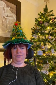 A phot of me from the shoulder up, I am beside a Christmas tree that has gold decorations and lights twinkling. I have a Christmas tree shaped hat on that is made of green tinsel and coloured Pom poms.