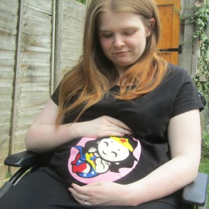 Ami is sitting in her wheelchair, she has long brown hair and is wearing a black t shirt with a baby superhero on the front. Ami has a bump and looks pregnant. 