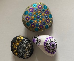 Three painted stones. One grey with blue and gold circle pattern. One black with gold and silver clusters of dots. One white with purple and black mandala designs. 