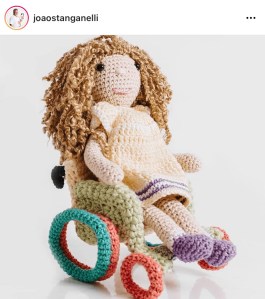 A crochet doll with brown curly hair in a crocheted green and orange wheelchair. 