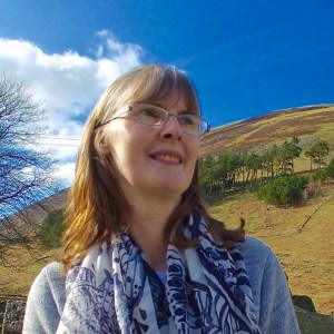 A portrait photo of Liz. She has light brown shoulder length hair and is wearing glasses. There is a blue sky and green hilly landscape in the background.