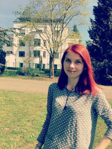 A photo of Caz from the waist up. She is standing and wearing a grey top. She has long red hair that is being slightly blown by the wind. In the background are trees, a large white building and blue sky. 