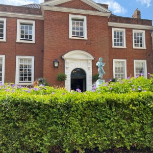 A green hedge is across the bottom. Above a Georgian style building can be seen. There is a water fountain coming up just above the hedge. The sky is bright blue. 