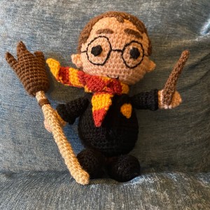A crochet Harry Potter toy. Holding a crochet wand and broomstick. He is wearing the Gryffindor scarf or red and yellow stripes.
