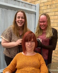 Me with red hair and an orange jumper sitting in my wheelchair. Two female friends are standing behind me. Both with long dark blonde hair. We are all smiling towards the camera.