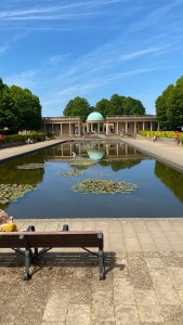 A photo looking across a square lake at a domed bandstand in the distance. The sky is blue. 