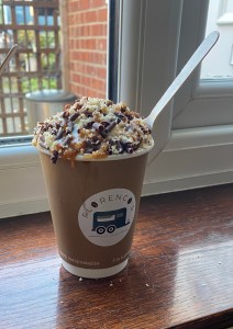 Hot chocolate in a brown cardboard cup with a wooden spoon sticking out. It is topped with cream, chocolate swirls, crumbled shortbread and caramel sauce. The cup is sitting on the inside of a windowsill.