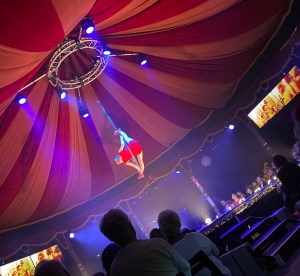 A lady in bright red gymnastics suit is hanging from a rope performing aerial acrobatics. Her legs are doing the splits along the rope that is hanging from the centre of canvas draped ceiling. 