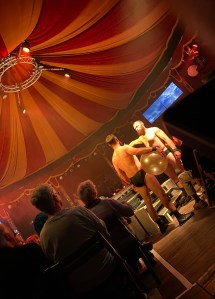 Two bearded white men wearing only black pants stand just off stage within the audience. They are facing each other and one is holding a large ball. 